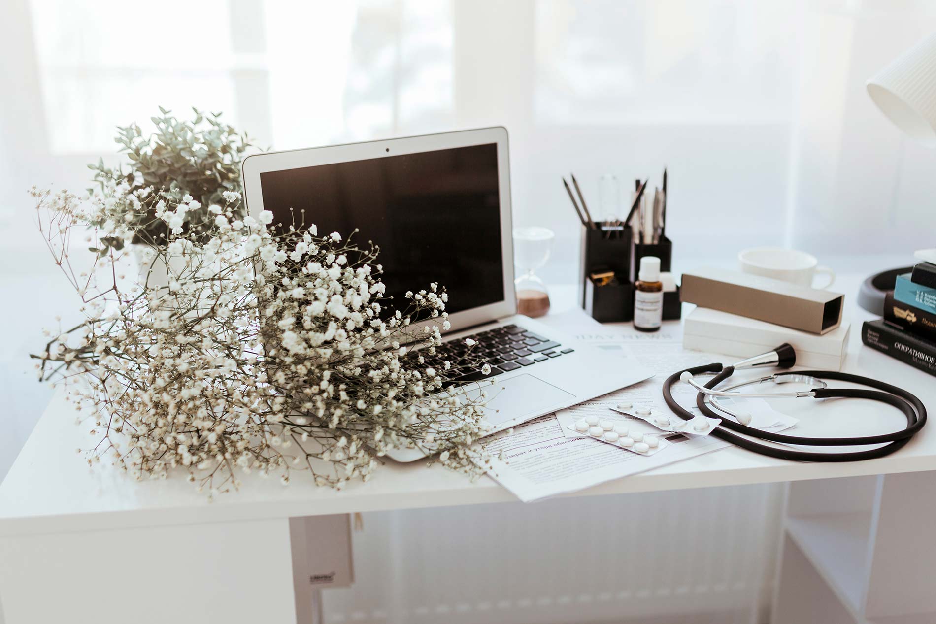 A bright, clean doctor’s desk with a laptop, flowers, and various medical items